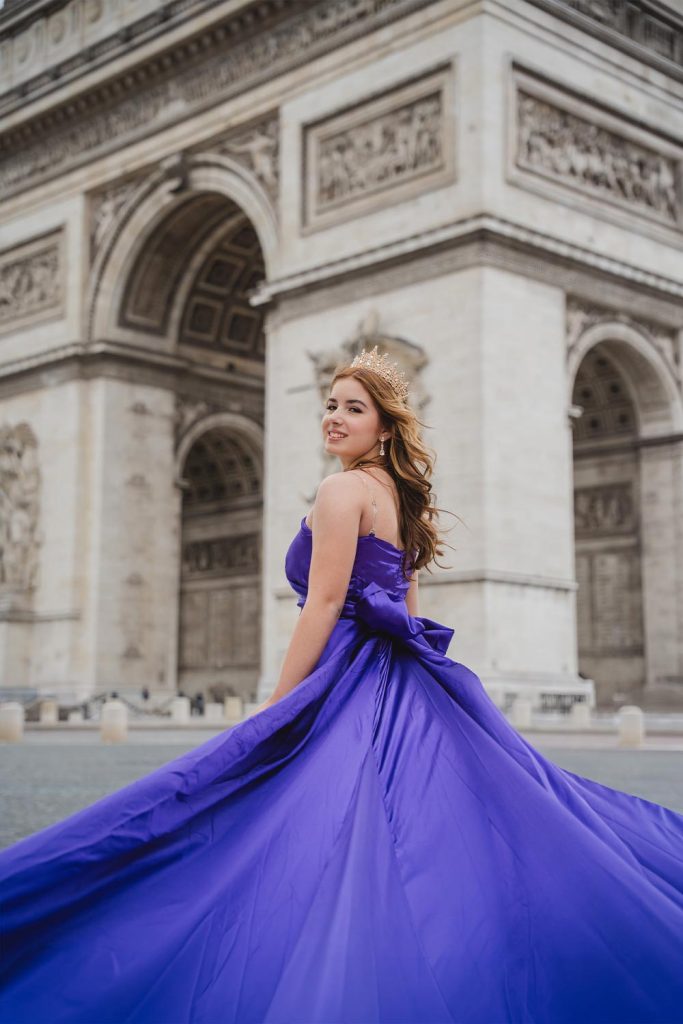 Quinceañera con vestido morado de cola larga y corona sonriendo frente al Arco de Triunfo en París / Quinceañera in a long-train purple gown and crown smiling in front of the Arc de Triomphe in Paris