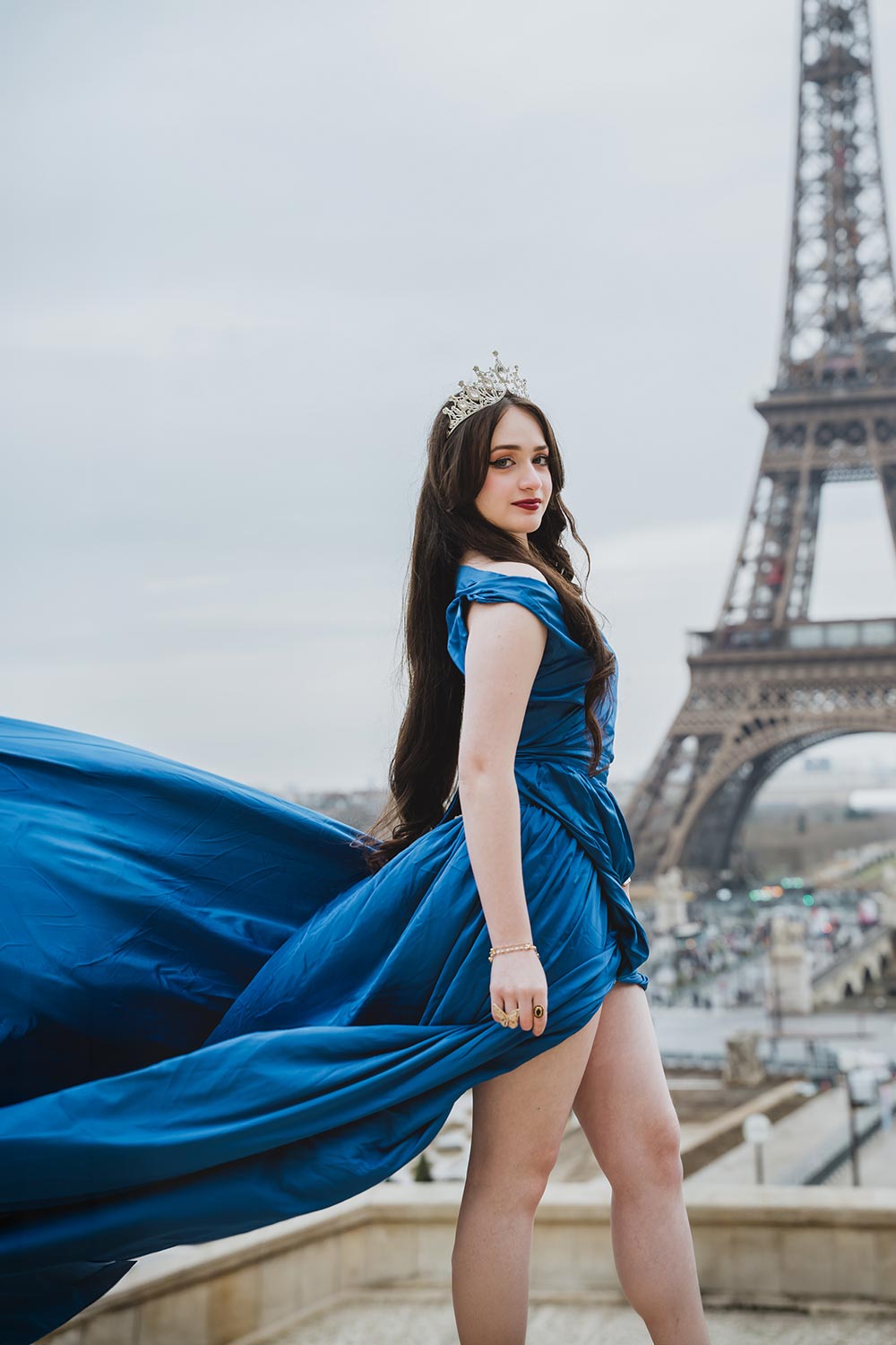 Quinceañera con vestido azul de tela voladora y corona posando frente a la Torre Eiffel desde el Trocadéro / Quinceañera in a flowing blue gown and crown posing in front of the Eiffel Tower at the Trocadéro