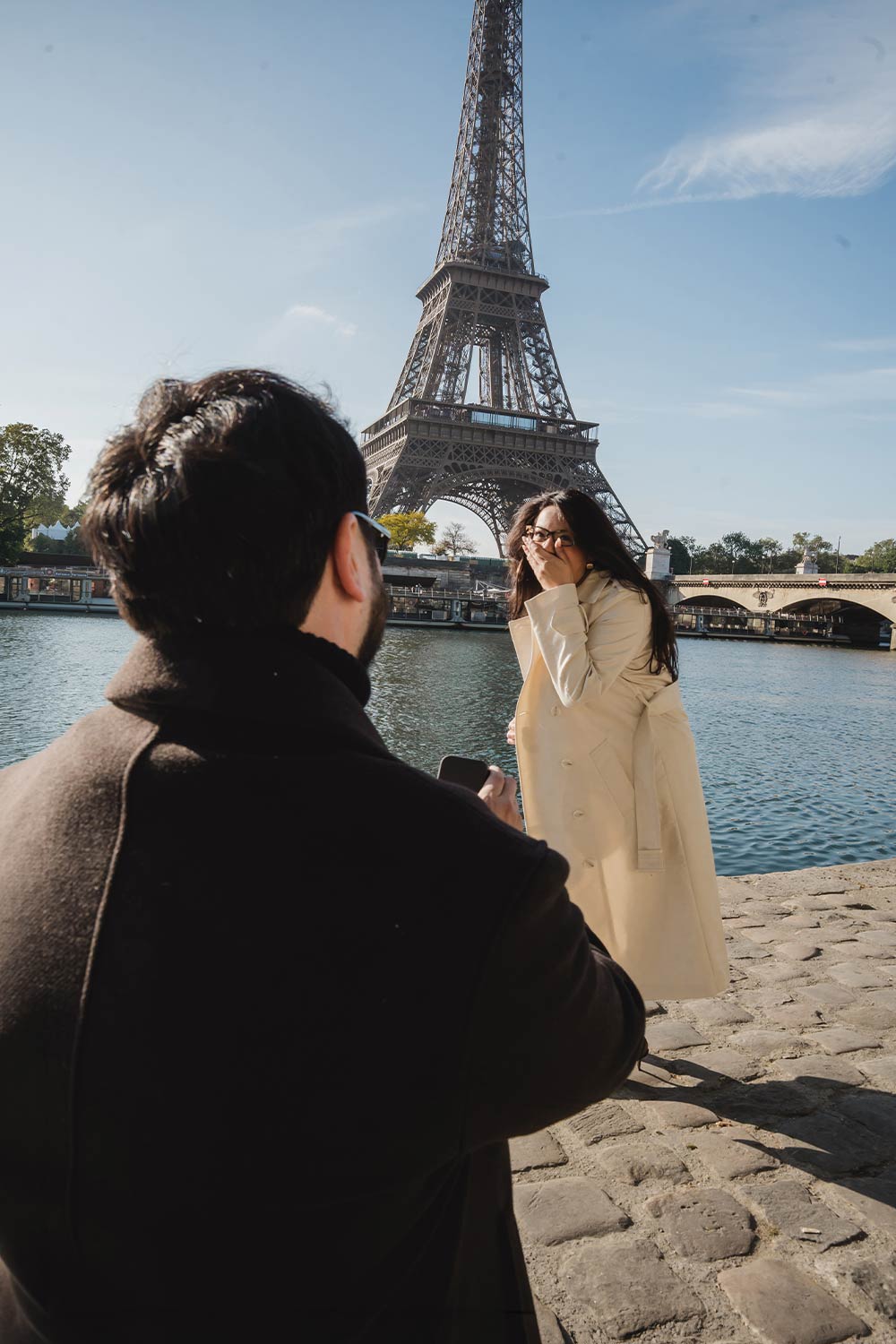 Hombre proponiendo matrimonio junto al río Sena mientras ella reacciona sorprendida con la Torre Eiffel de fondo en París / Man proposing marriage by the Seine river as she reacts in surprise, with the Eiffel Tower behind, Paris