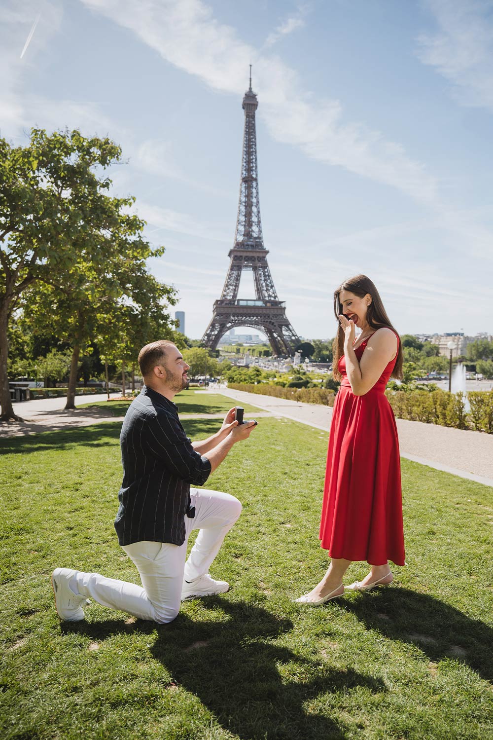 Hombre proponiendo matrimonio de rodillas en los jardines del Trocadéro mientras ella con vestido rojo reacciona sorprendida, Torre Eiffel de fondo / Man kneeling to propose marriage in the Trocadéro gardens as she reacts in surprise wearing a red dress, Eiffel Tower behind
