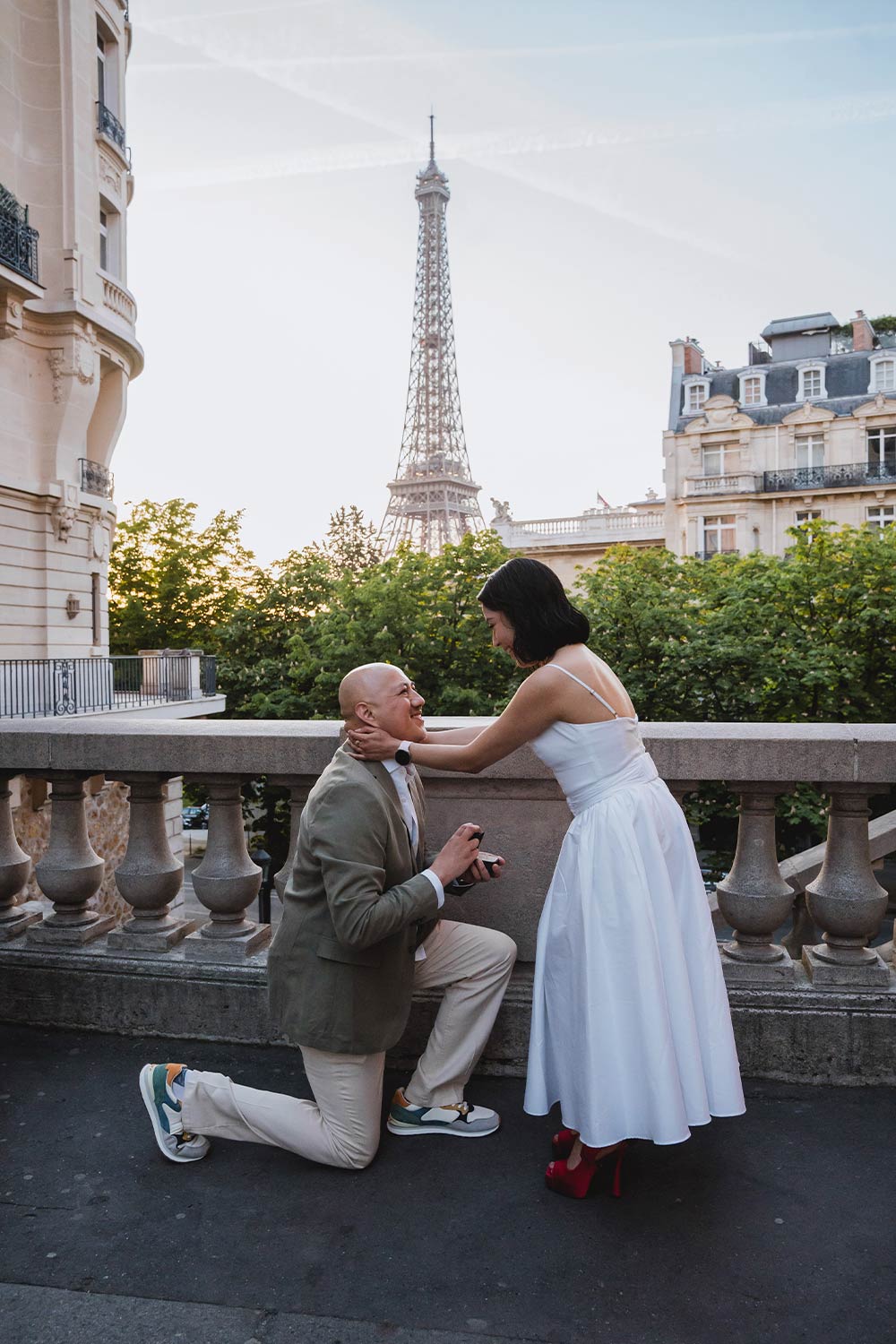 Hombre proponiendo matrimonio de rodillas en un balcón parisino al atardecer, ella con vestido blanco, Torre Eiffel de fondo / Man kneeling to propose marriage on a Parisian balcony at sunset, her in a white dress, Eiffel Tower behind