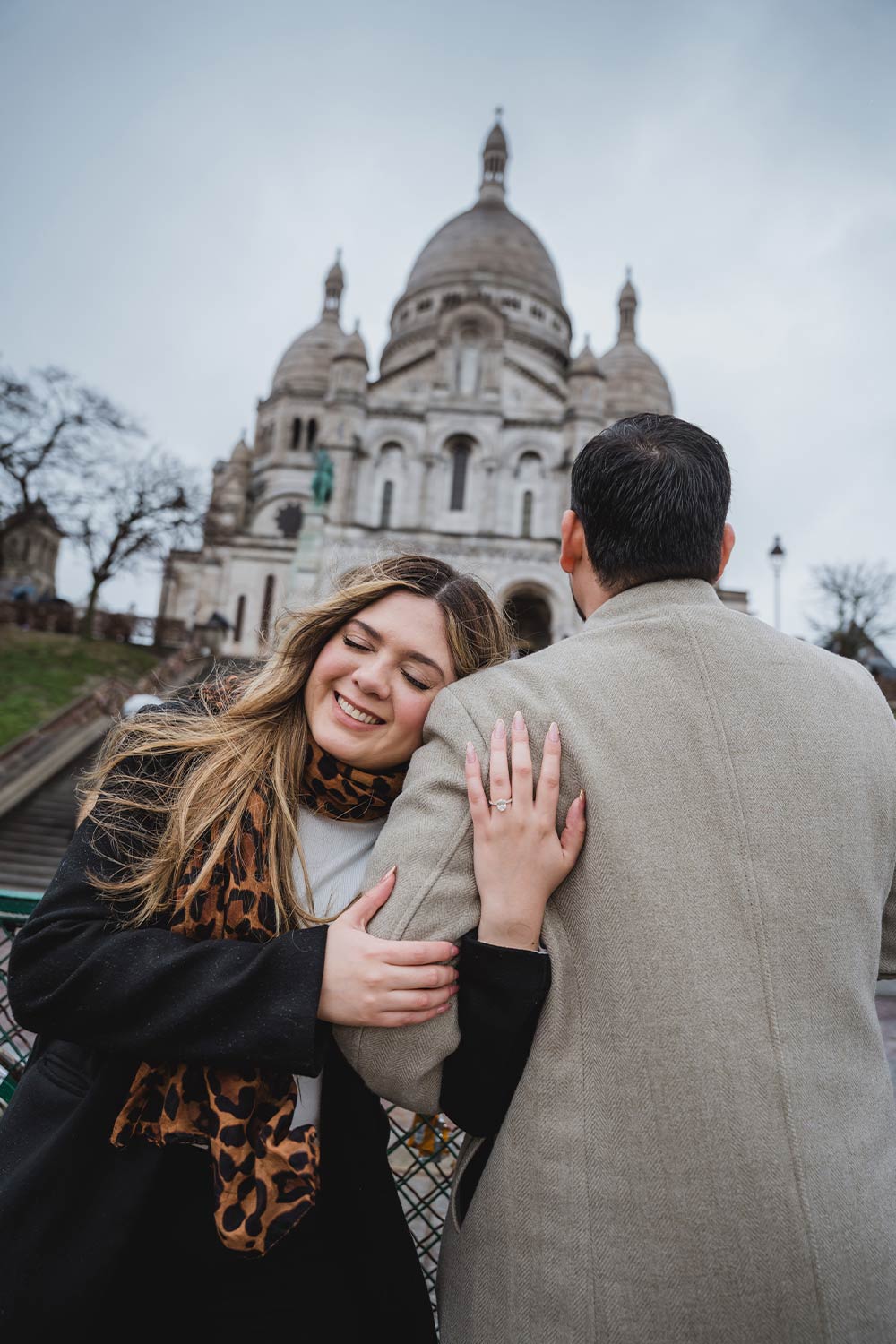 Mujer abrazada a su pareja mostrando el anillo de compromiso frente a las escaleras del Sacré-Cœur en Montmartre / Woman embracing her partner showing her engagement ring in front of the Sacré-Cœur stairs in Montmartre